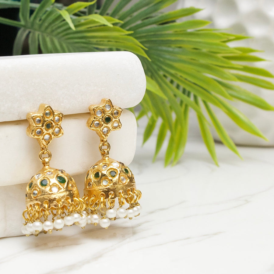 Gold-plated jhumka earrings featuring green gemstone accents, floral studs, and pearl dangles displayed on white marble with green foliage background