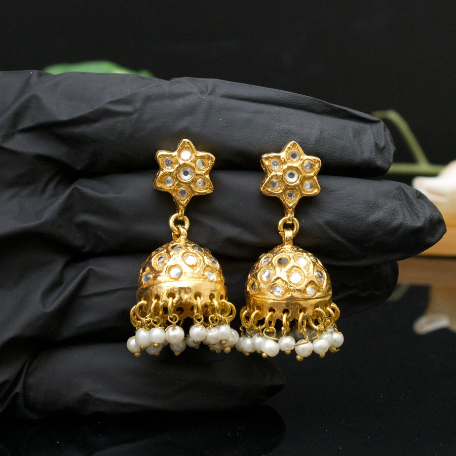 Hand holding gold-plated jhumka earrings with clear gemstone floral studs and pearl dangles shown from front view on black background