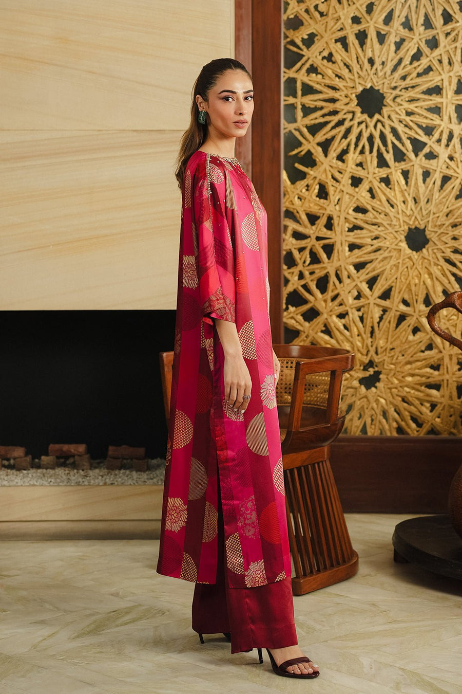 Side profile of a woman modeling a long red and burgundy silk dress with gold detailed circles and floral motifs, showing the loose silhouette and side slit, paired with burgundy pants and black heels.