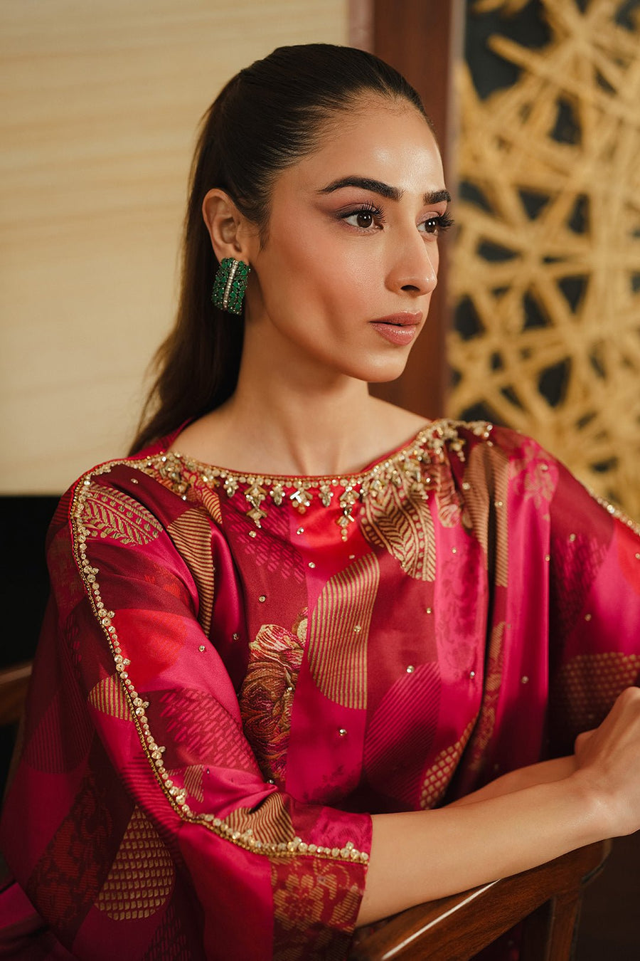 Close-up of the embellished neckline and three-quarter sleeve border of a red charmeuse silk dress featuring gold beadwork and intricate pattern details worn by a woman with pulled-back hair and green earrings.