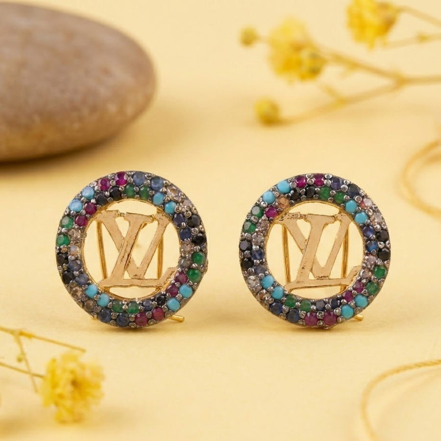 Close-up of gold-tone round earrings with multicolor stones arranged on a yellow surface with dried flowers