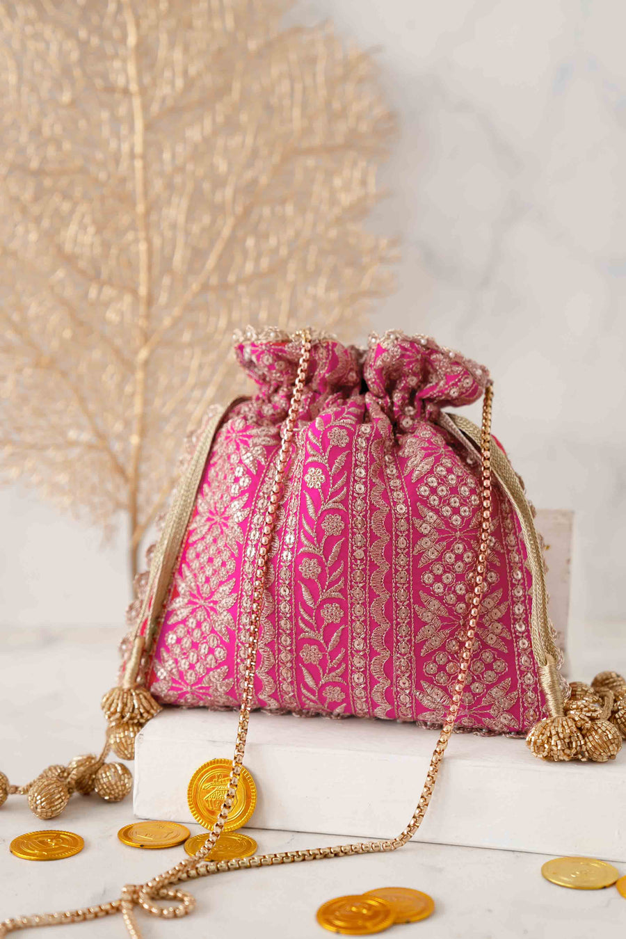 Pink potli bag with gold floral embroidery, gold drawstring tassels, and gold chain sling displayed on a white block with gold coins scattered around