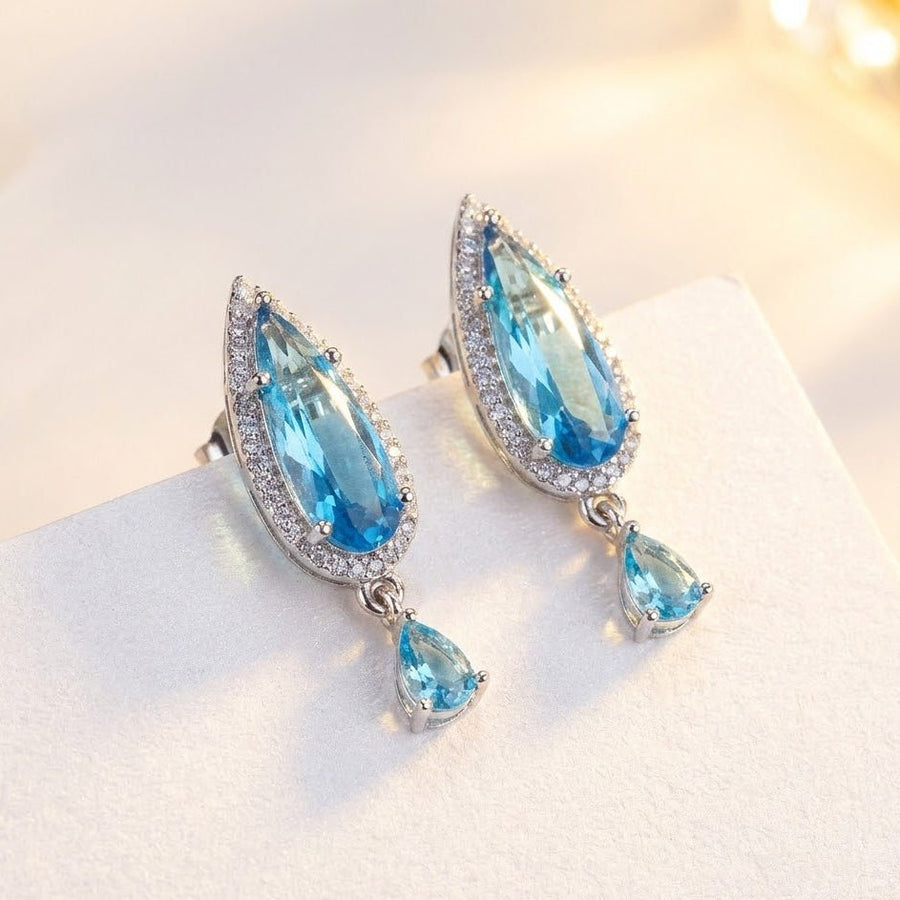 Blue teardrop crystal earrings with halo of clear stones and small dangling crystal resting on light surface
