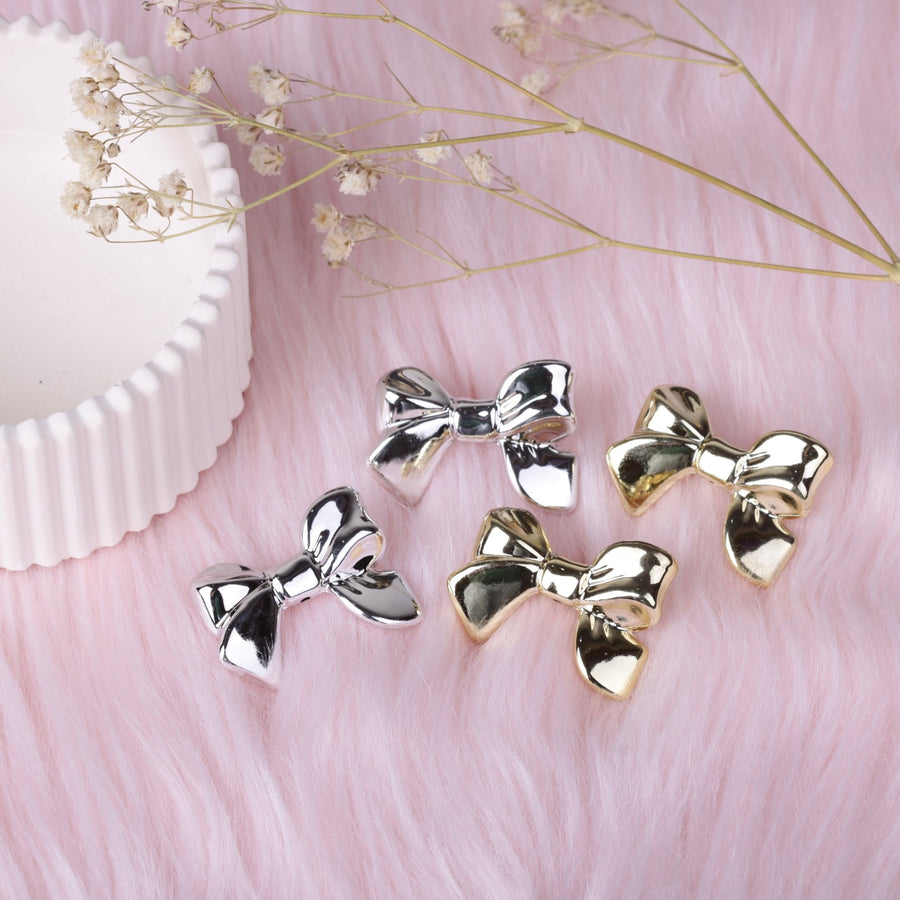 Two pairs each of polished gold and silver bow stud earrings arranged on soft pink faux fur background with dried flowers nearby