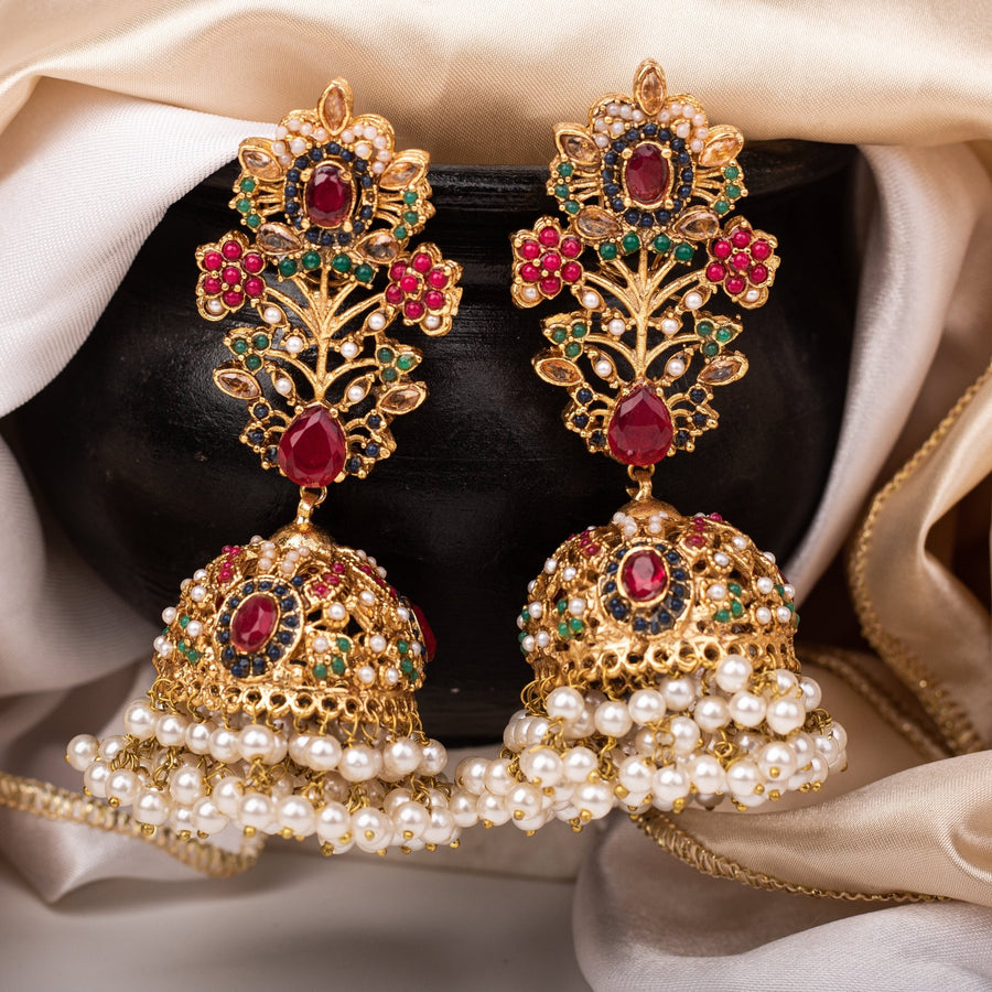 Pair of Royal Garden Jhumka earrings with deep red gemstones surrounded by multicolored stones and pearl dangles displayed on a black pot with beige fabric backdrop