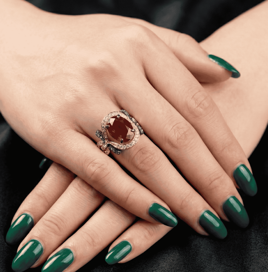 Rose gold ring on model's hand showcasing a large oval red-brown gemstone with halo stones and black oxidized bow detailing