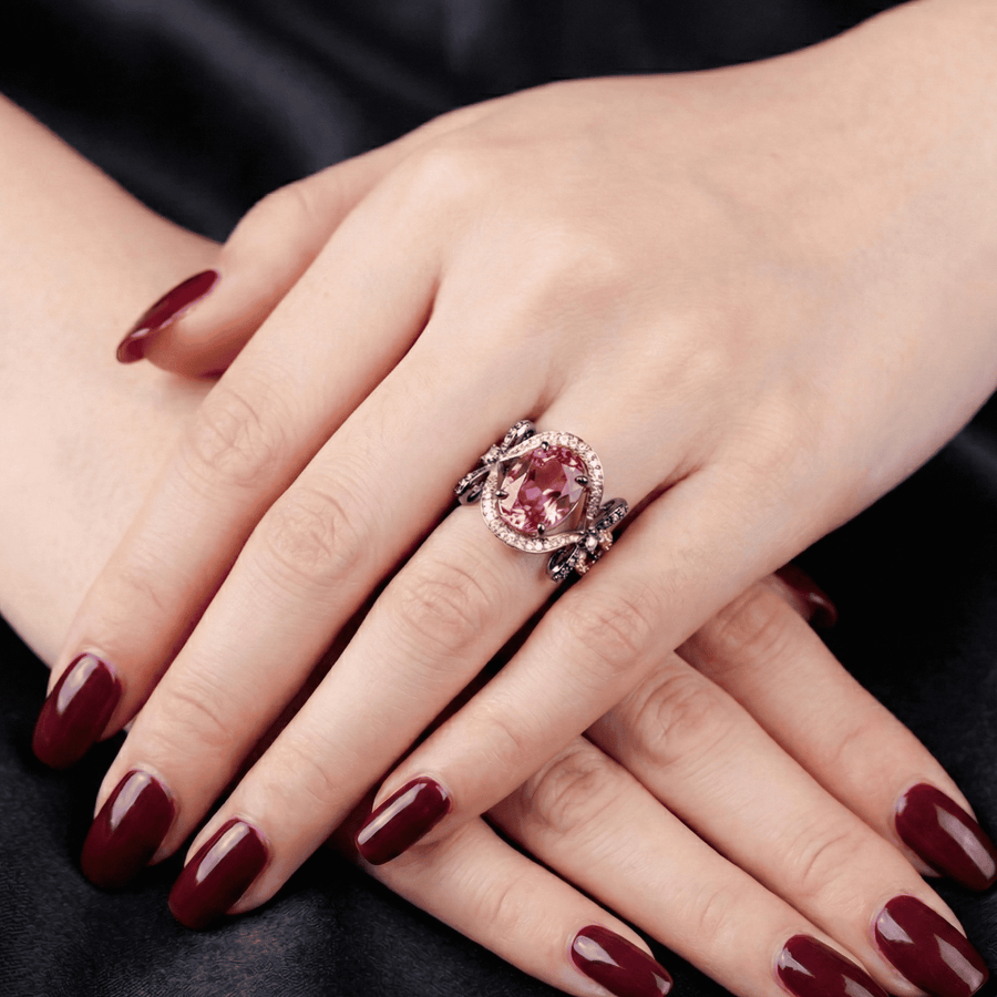 Model wearing rose gold ring with a large oval pink gemstone surrounded by a halo of small clear stones and ornate black oxidized bow details