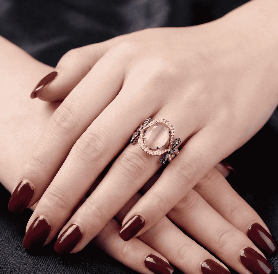 Hand adornment with rose gold ring showcasing a large deep pink semi-precious oval gemstone framed by sparkling clear stones and black oxidized bows