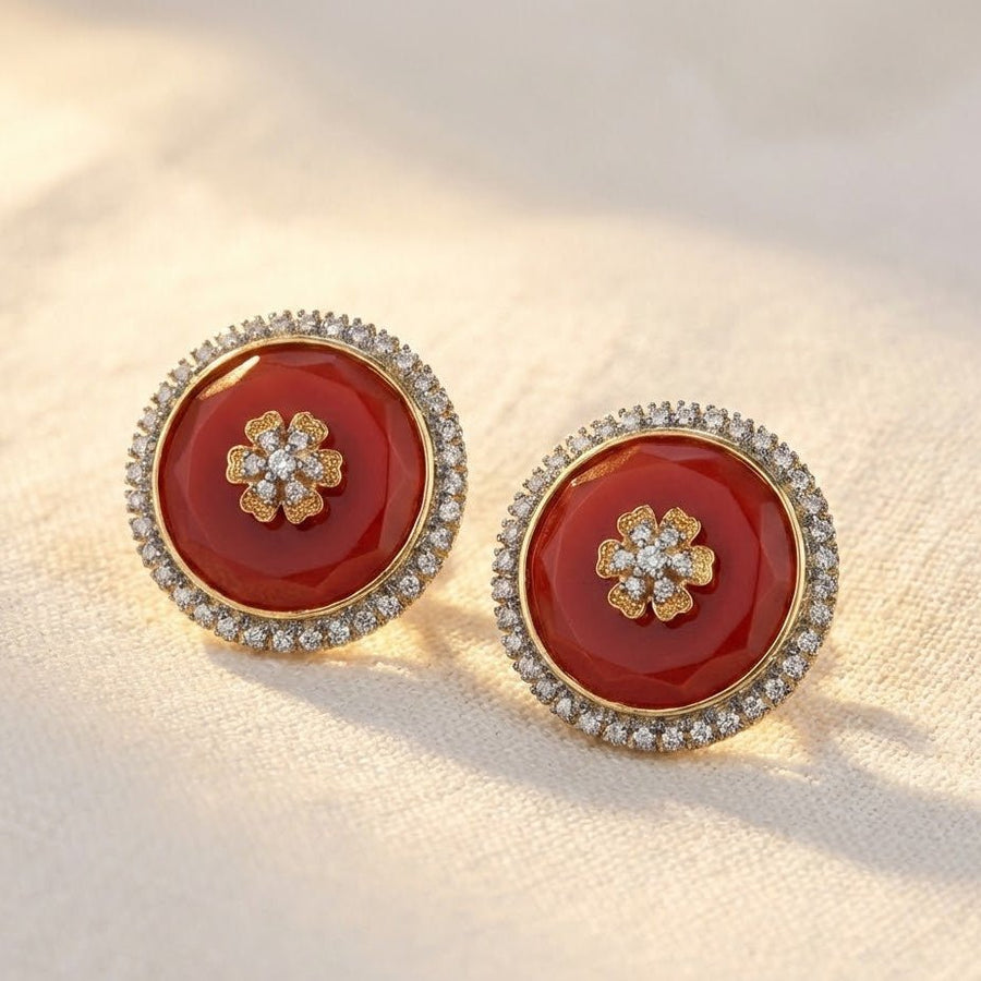 Round stud earrings with red faceted stones and sparkling halos placed on light fabric with natural yellow flowers
