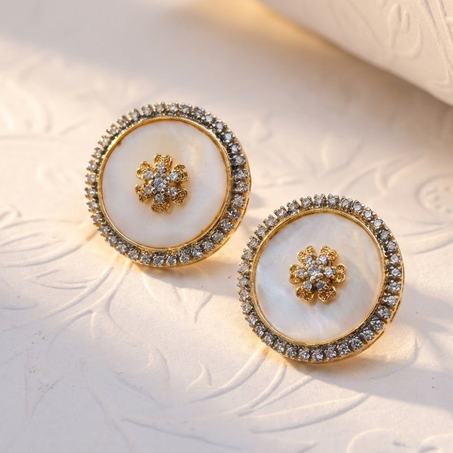 White mother of pearl round stud earrings with flower motif and sparkling stone halo displayed on embossed light surface