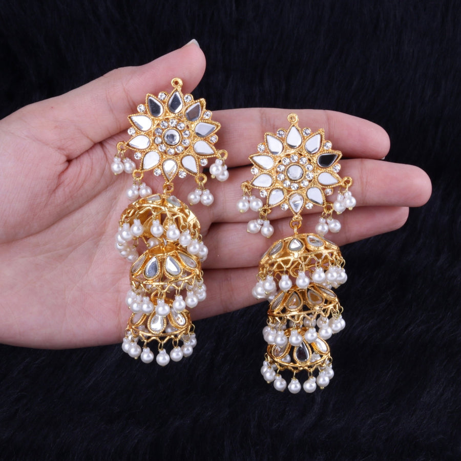 Close-up of a pair of gold-tone chandbali earrings with mirror embellishments and cascading white faux pearls held in a hand against a black textured background