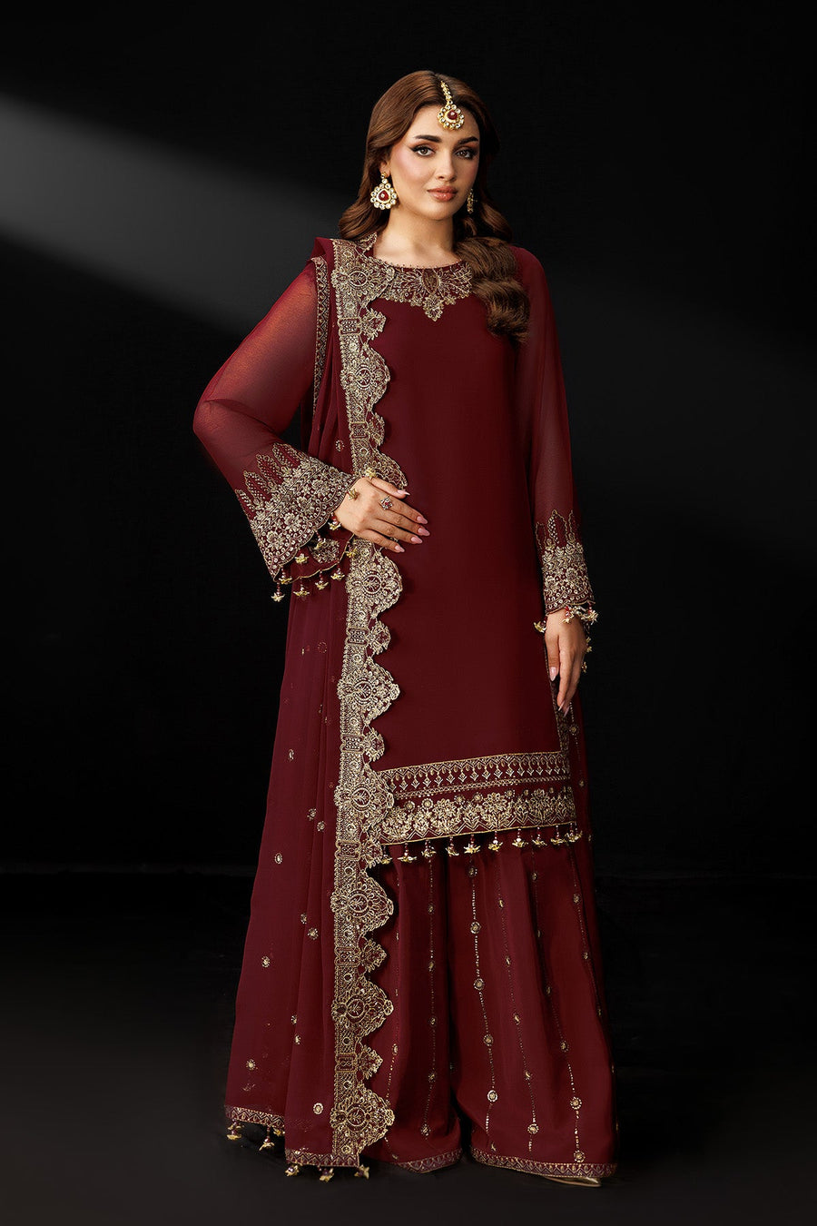 Woman wearing maroon traditional embroidered suit with sheer sleeves and embellished dupatta against black background