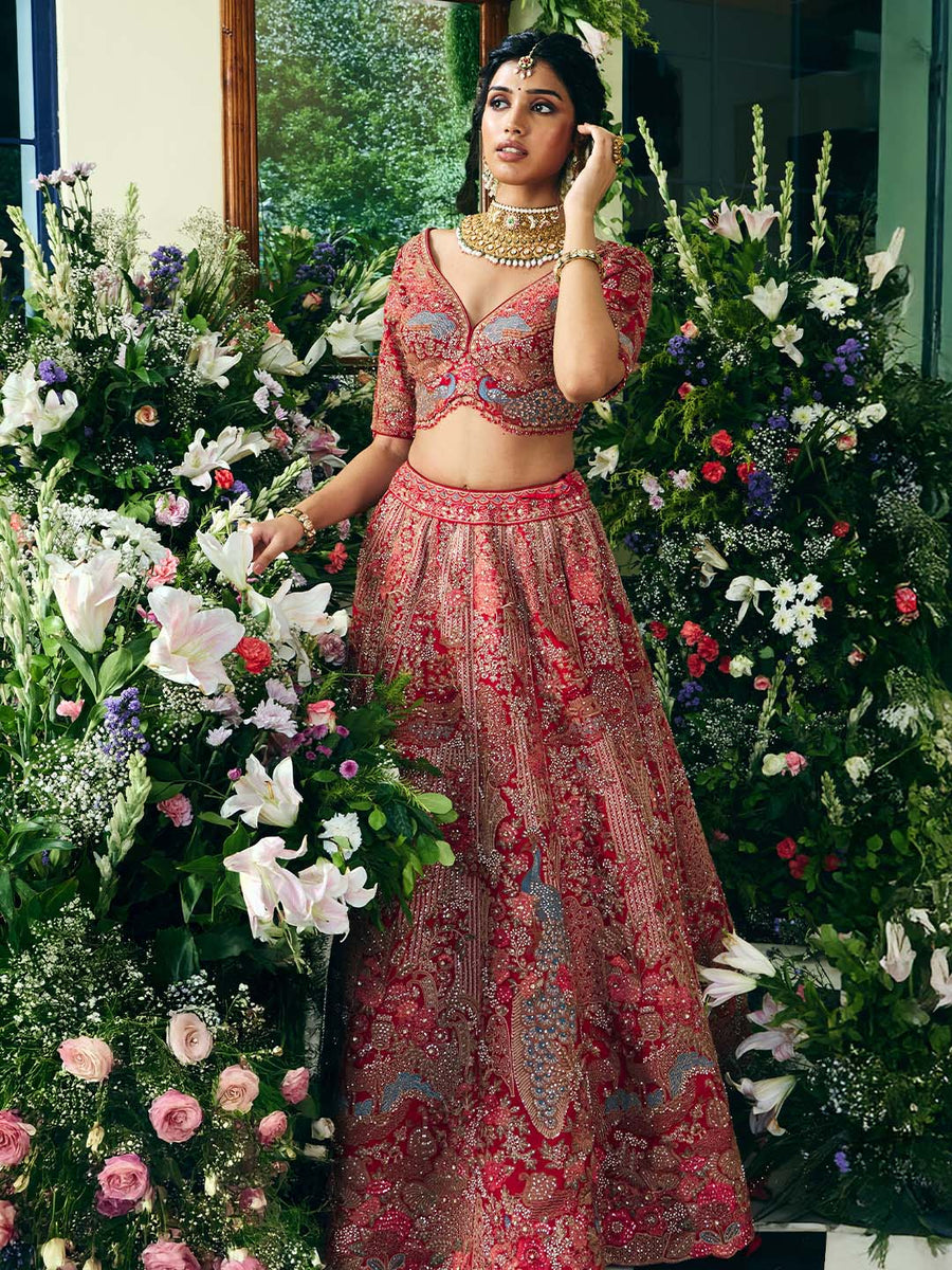 Model displaying the full red lehenga skirt with detailed embroidery and a matching embellished blouse, surrounded by flowers.
