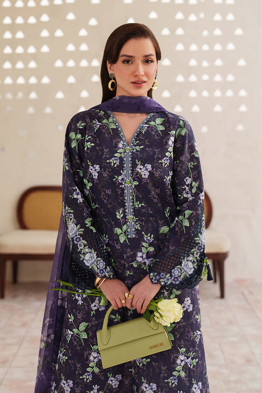 Side profile of woman in navy floral lawn suit with sheer dupatta flowing behind her, white floral pants, and gold heels, standing near a cream column.