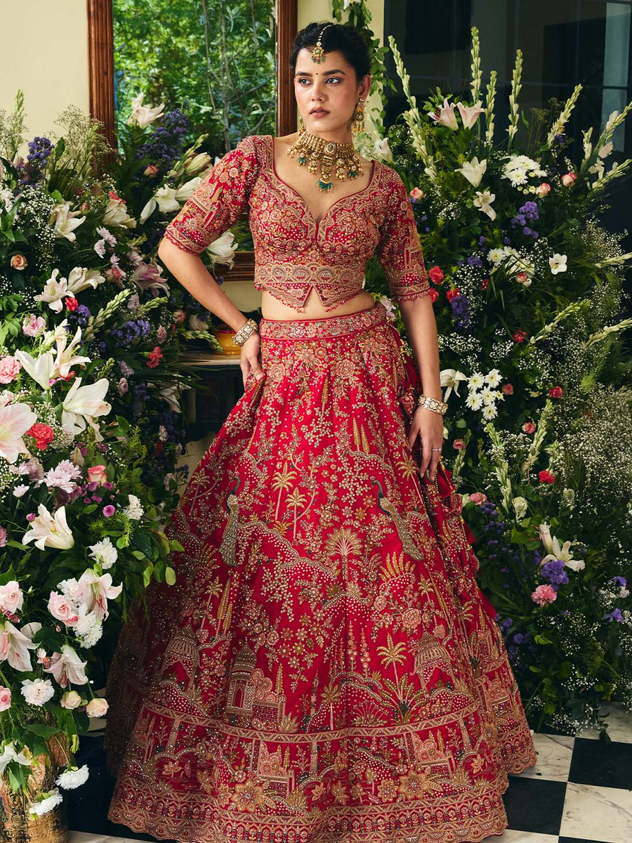 Model wearing a luxury red bridal lehenga with intricate gold embroidery standing among lush floral arrangements