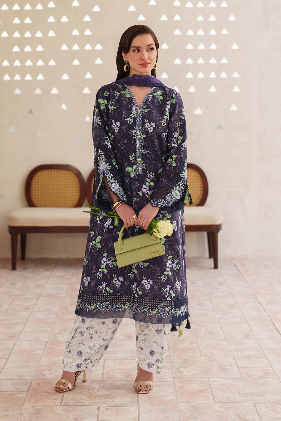 Woman standing wearing a navy floral embroidered lawn suit with white floral pants and holding a pale green handbag and white roses, posing against a textured neutral wall with wooden chairs.