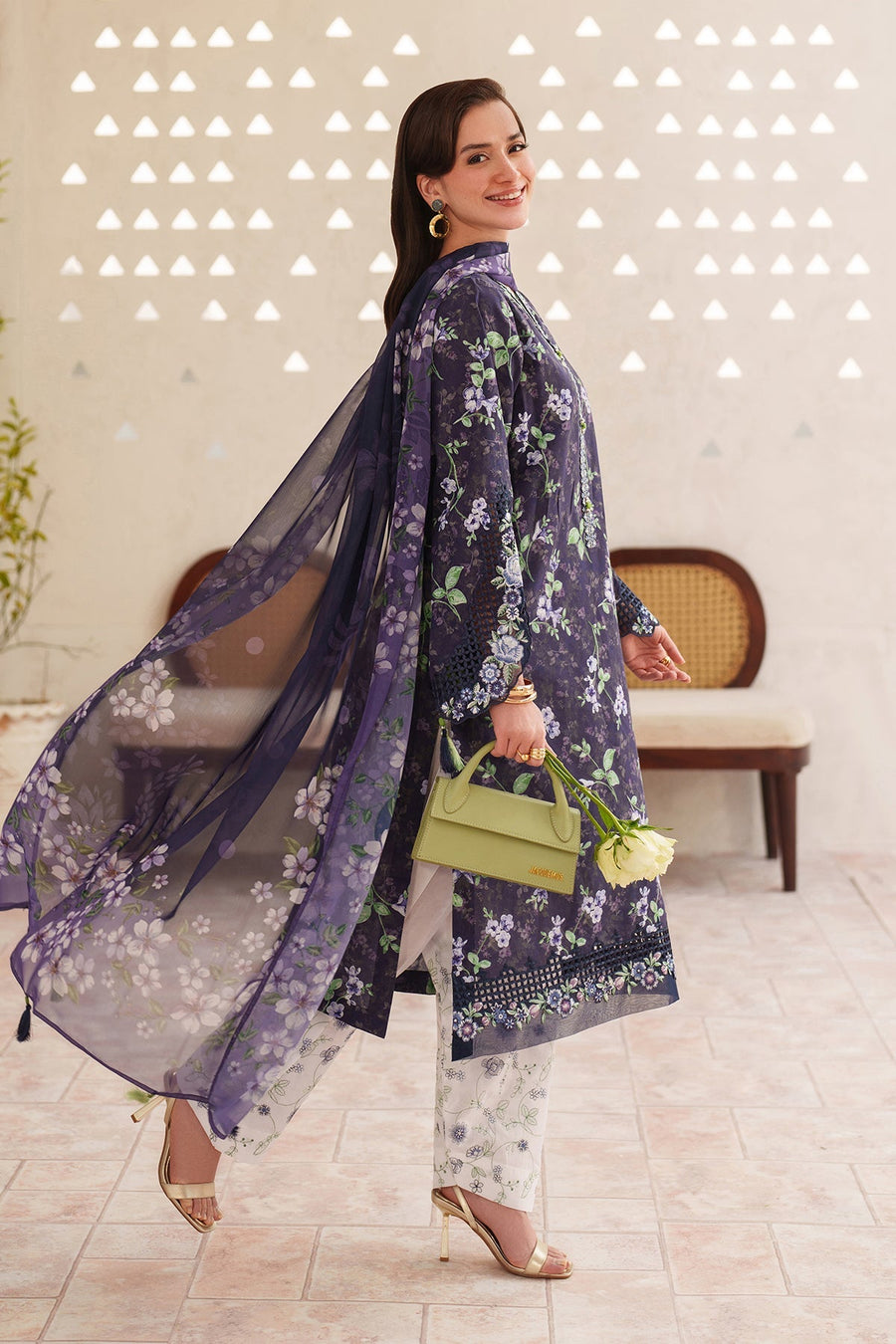 Woman posing against a wall wearing a navy floral embroidered kurta, white floral pants, holding a pale green handbag and white roses, smiling with wavy hair.