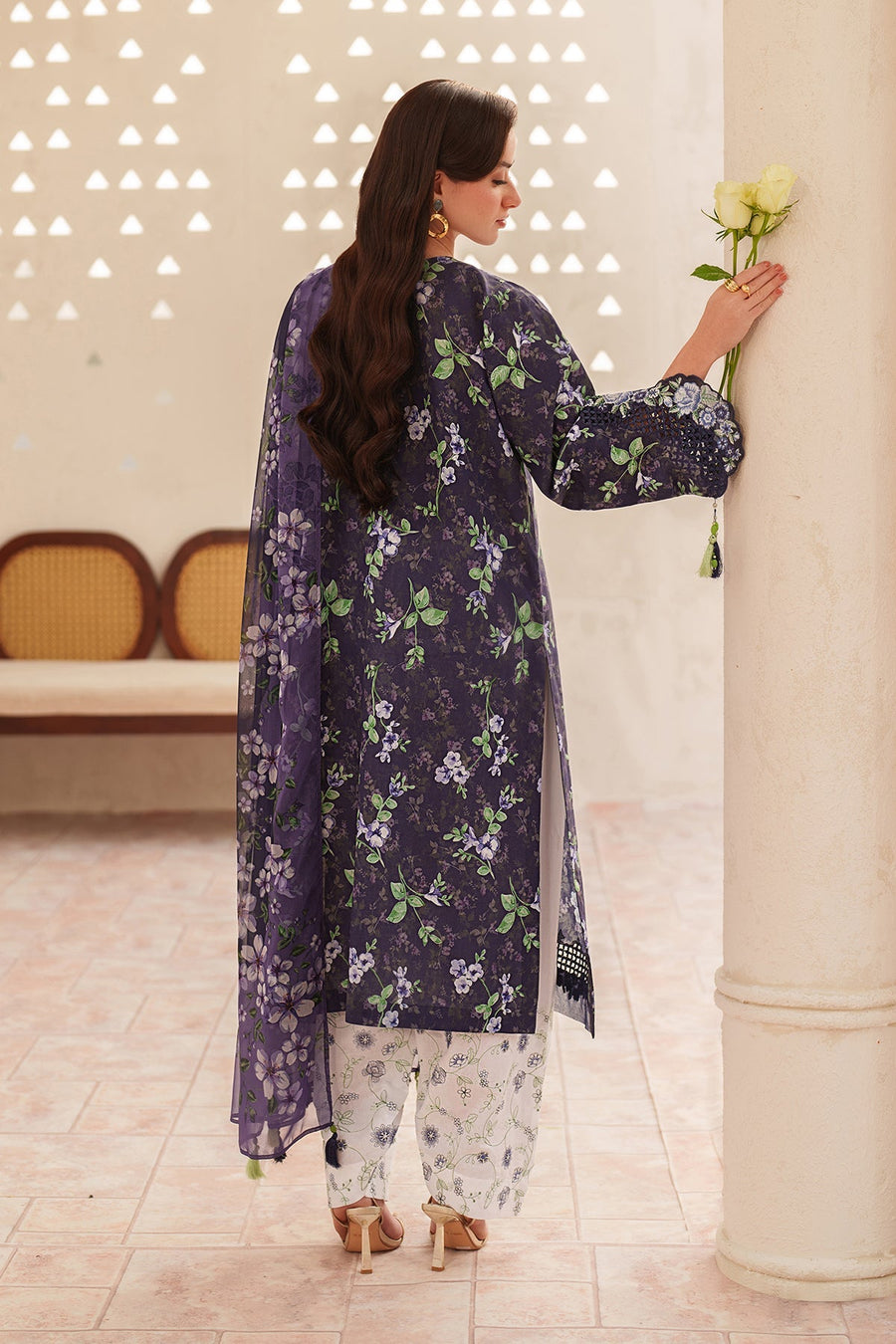 Back view of woman in floral embroidered navy kurta with matching sheer navy dupatta and white printed pants, holding white roses, standing near a column.