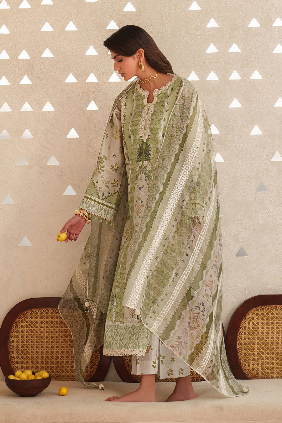 Side view of woman wearing embroidered lawn suit and dupatta, holding small yellow lemons in hand, adorned with gold jewelry