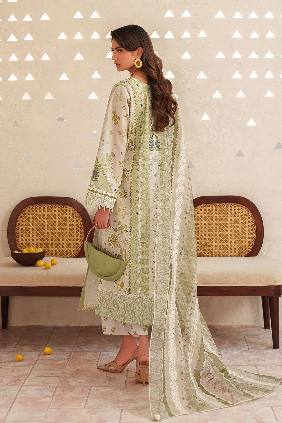 Back view of woman dressed in green and white floral printed embroidered lawn suit, carrying a green handbag with gold handle, standing near rattan bench