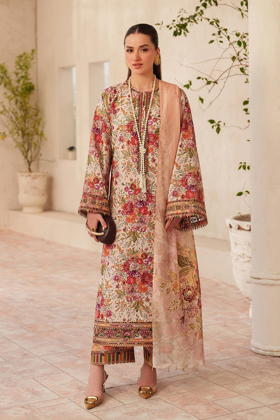 Model wearing a floral embroidered lawn kameez with peach embroidered dupatta and golden shoes, holding a brown clutch