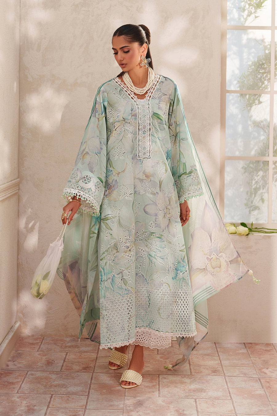 Woman wearing a pastel green embroidered lawn suit with floral designs, holding a sheer white handbag filled with white roses, standing indoors on tiled floor
