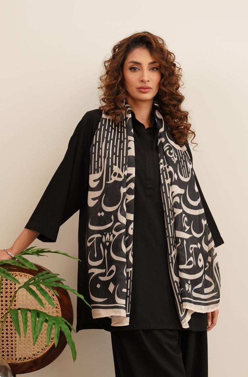 Woman wearing a black and cream scarf with intricate Arabic calligraphy design over a black outfit.