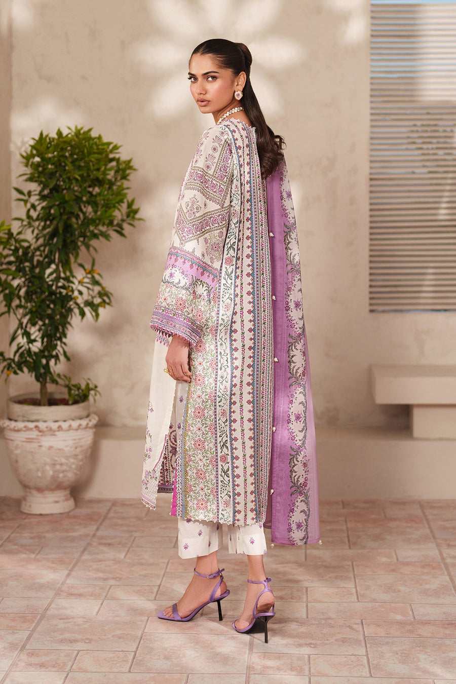 Side view of woman showcasing detailed embroidery on lawn suit with organza sleeves, holding white roses, styled with purple strappy heels.