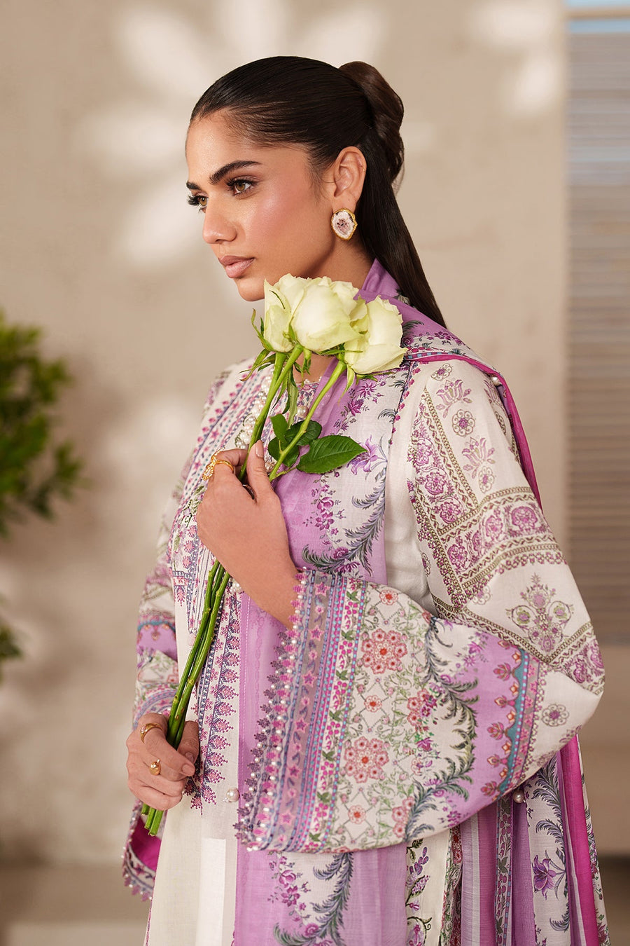 Close-up of detailed floral embroidery on cream lawn fabric showing fine stitches in lavender and pink, paired with matching trousers and white roses.