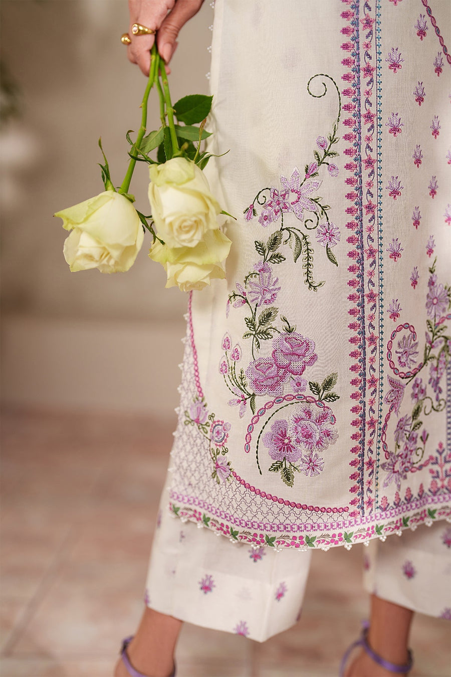 Portrait of woman holding white roses, highlighting embroidered neckline and organza sleeves with pearl embellishments on pastel floral lawn suit.