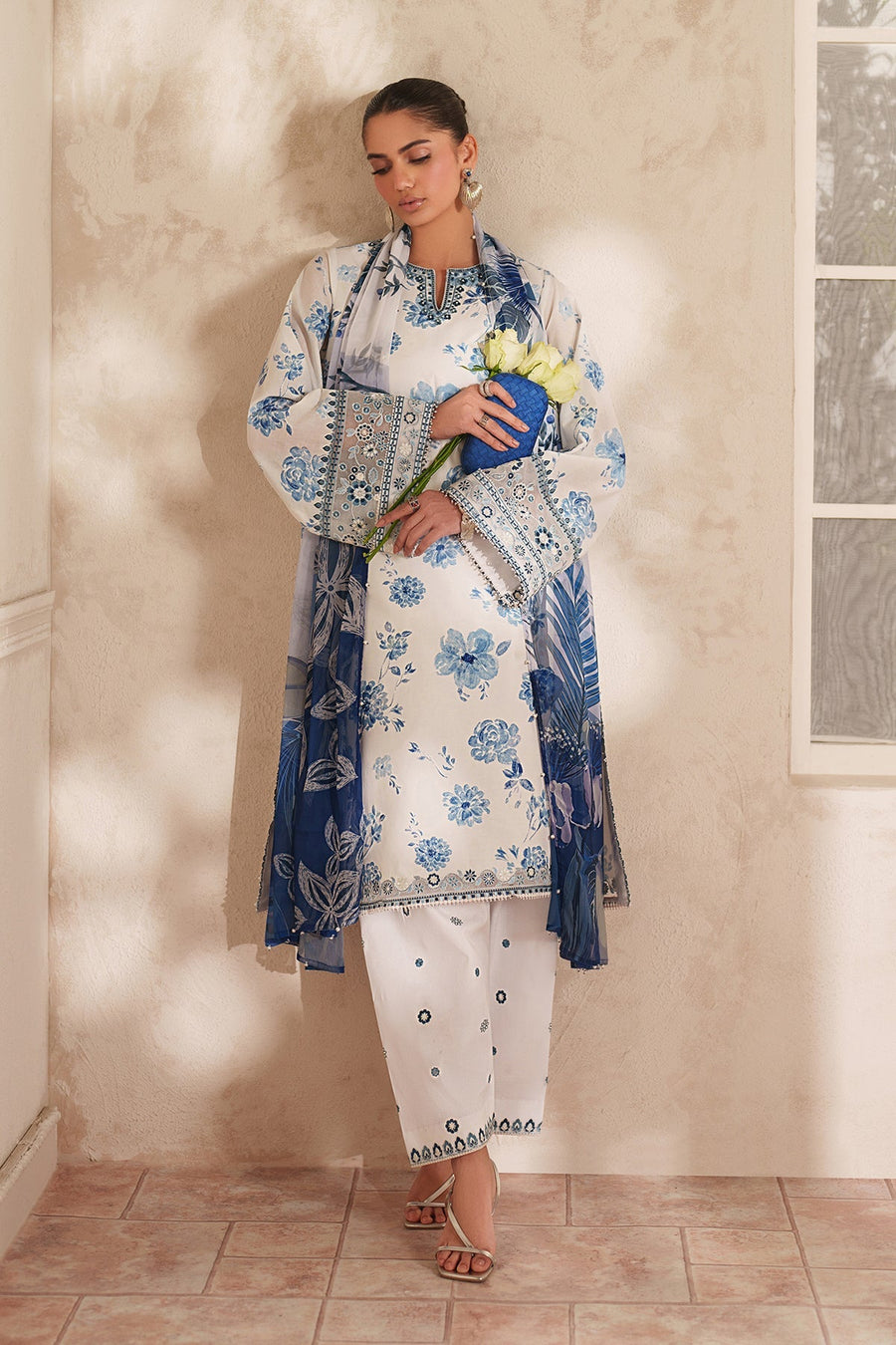 Side angle close-up of woman’s face showing large silver heart-shaped earrings and smooth hair bun, wearing embroidered blue floral lawn outfit.