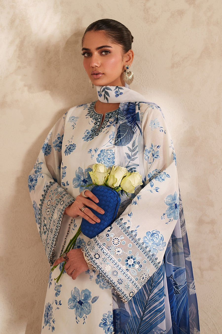 Close-up of model holding white roses and blue clutch, highlighting embroidered sleeve featuring floral patterns and bead accents