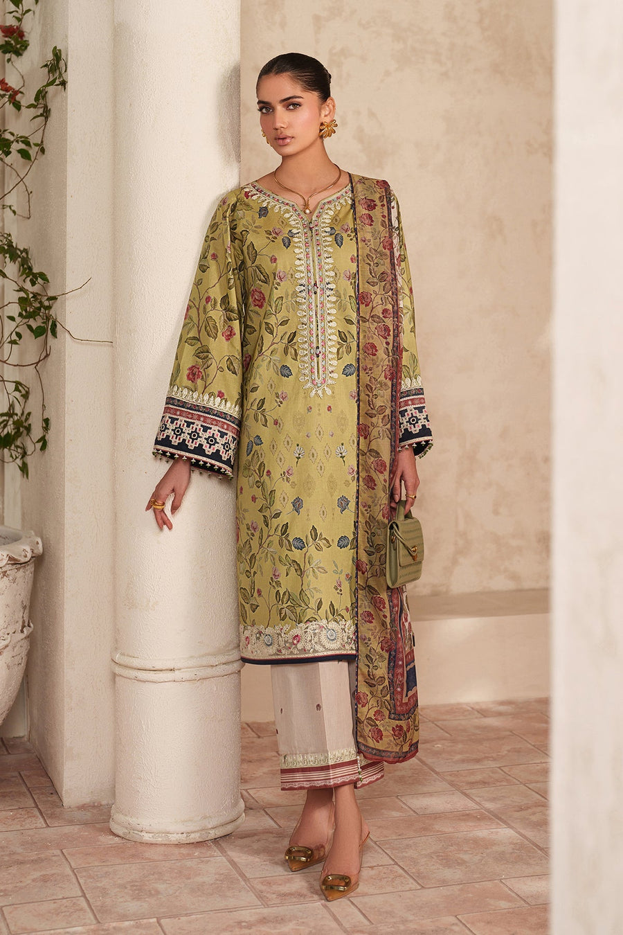 Woman standing against a column wearing a soft green floral embroidered lawn shirt with white wide-legged pants and holding a small green handbag
