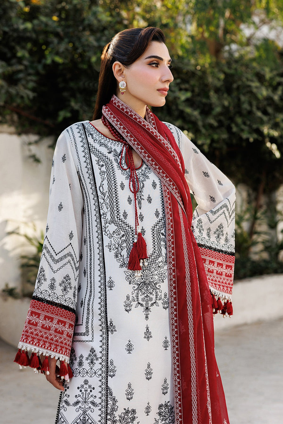Woman posing with embroidered lawn suit, holding red embroidered dupatta with tassels