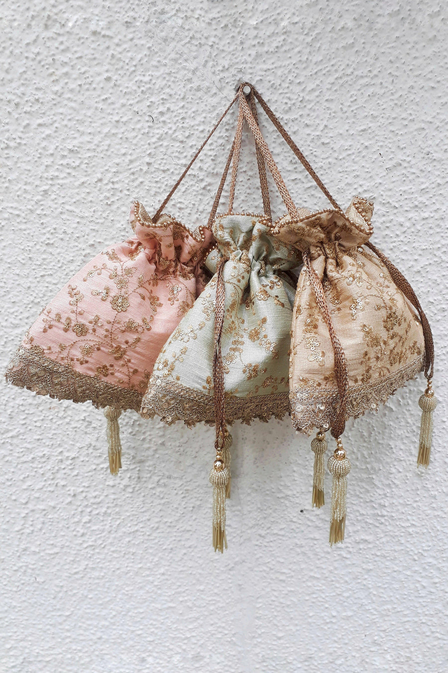 Set of three pastel silk potli bags with exquisite gold floral embroidery, drawstring closure, and tassel accents displayed against a textured white wall.