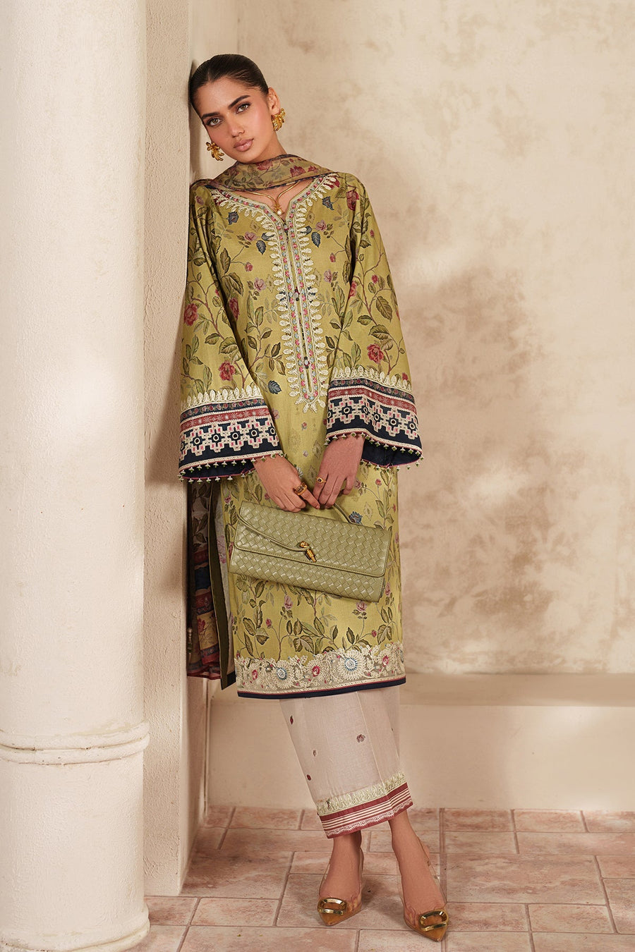 Woman facing slightly sideways wearing soft green floral embroidered lawn suit with matching dupatta, accessorized with statement gold earrings and rings