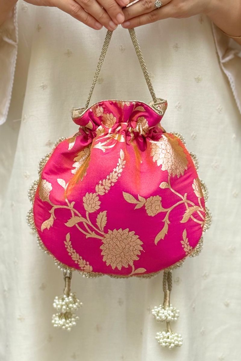 Rose pink Banarasi silk potli bag with gold floral embroidery and pearl tassels held against a cream embroidered fabric background