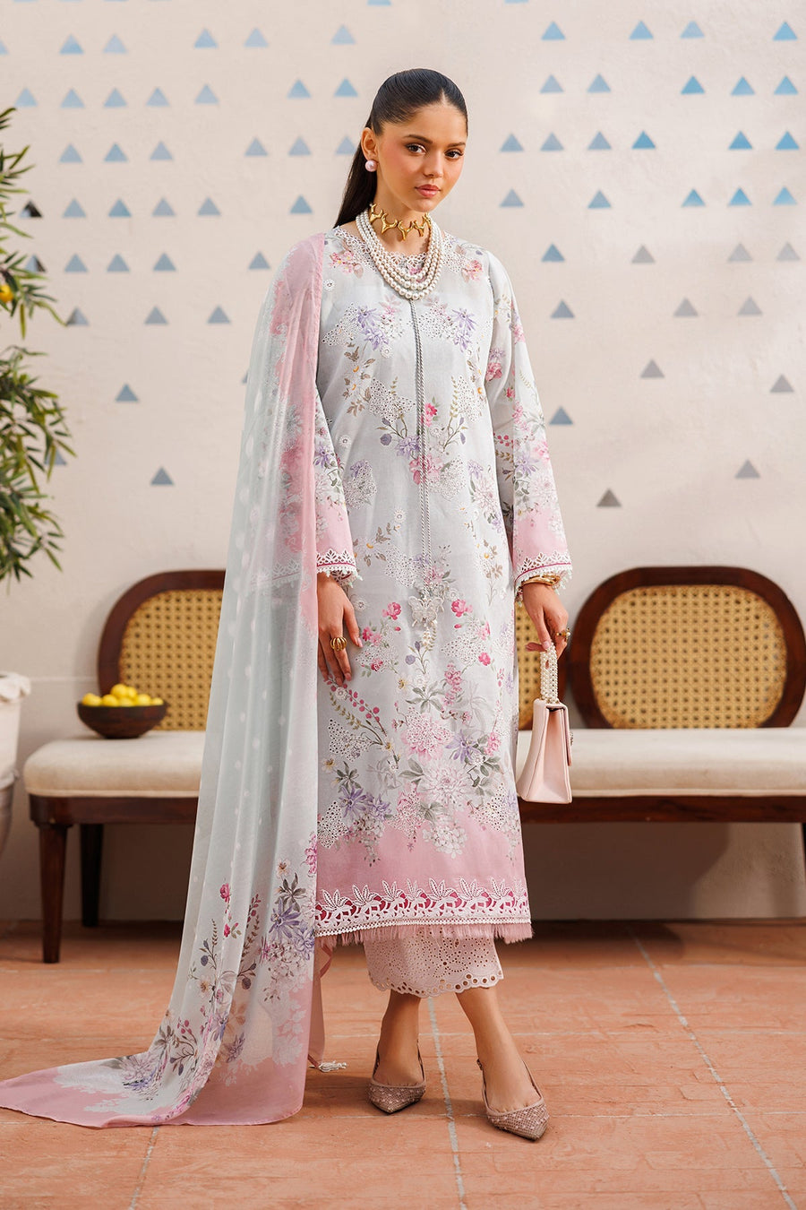 Front view of woman wearing floral embroidered lawn suit in pastel pink and lilac with organza sleeves and matching dupatta, holding a blush pink handbag