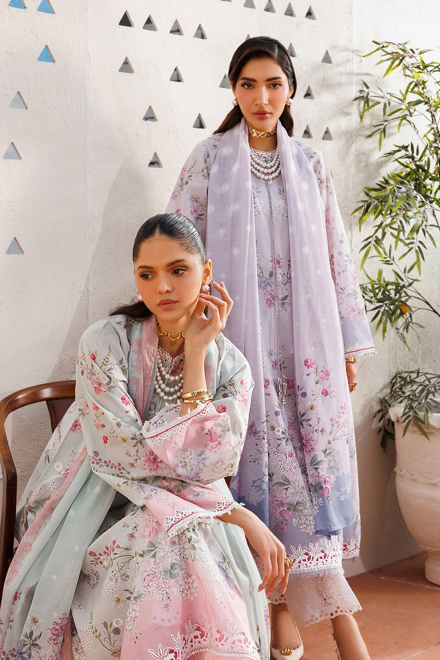 Two women styled in pastel floral embroidered lawn suits sitting on a bench, highlighting variety and complementary designs.