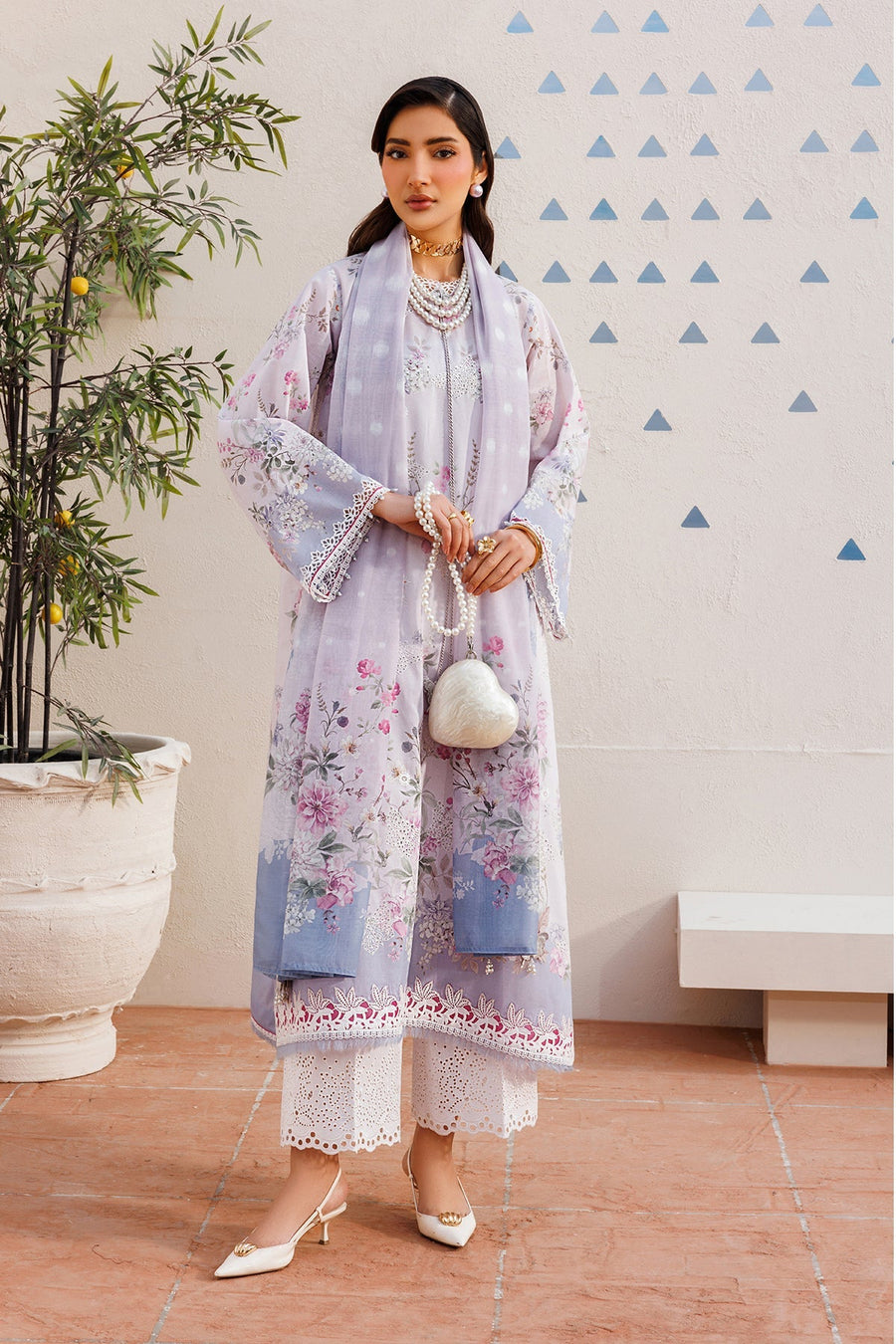 Woman wearing floral embroidered lavender lawn suit with white eyelet lace pants and matching dupatta, accessorized with pearl jewelry and heart-shaped purse, standing by a potted plant.