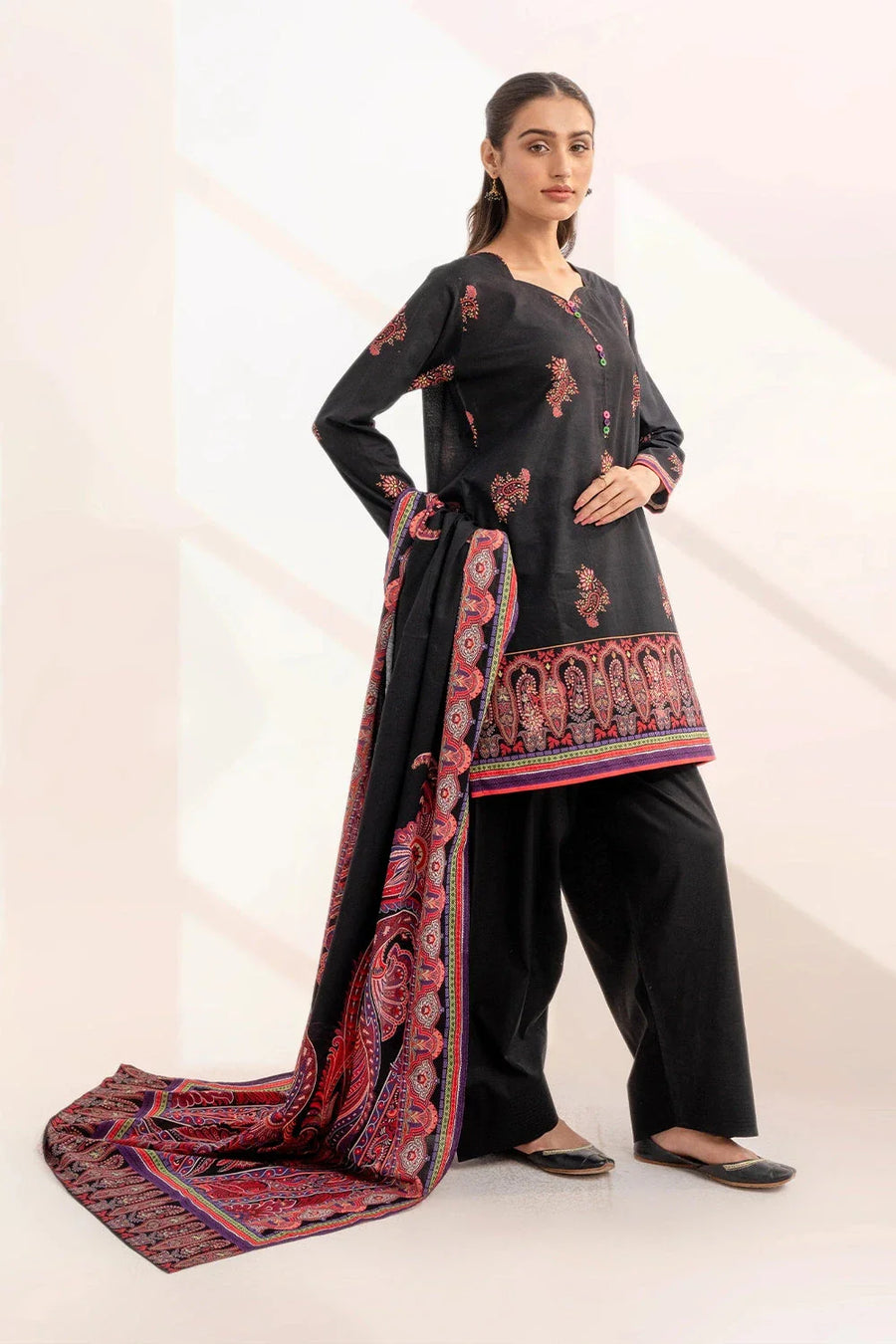 Woman posing in black printed three-piece lawn suit with dupatta draped over arm and traditional black footwear