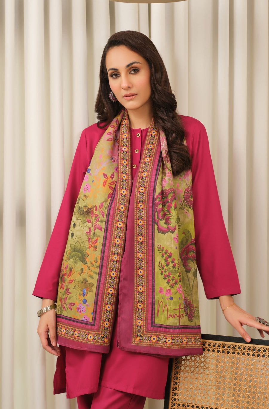 Woman wearing vibrant floral printed silk scarf over deep pink outfit, standing by woven chair in neutral background