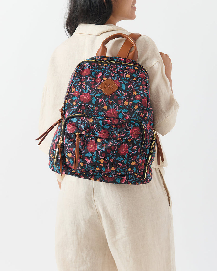 Woman wearing a floral vines printed backpack with brown leather accents and multiple zippered pockets, styled with a light beige outfit