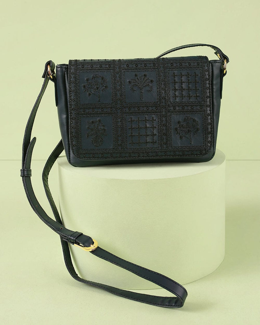 Black crossbody bag with detailed black embroidery on front flap, gold-tone hardware, and adjustable strap displayed on cream pedestal