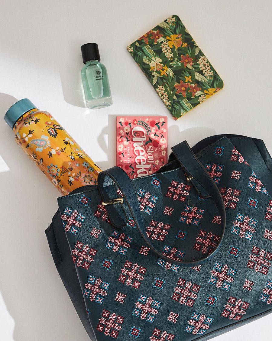 Flat lay of embroidered vegan leather tote bag surrounded by a floral water bottle, perfume, floral notebook, and face mask