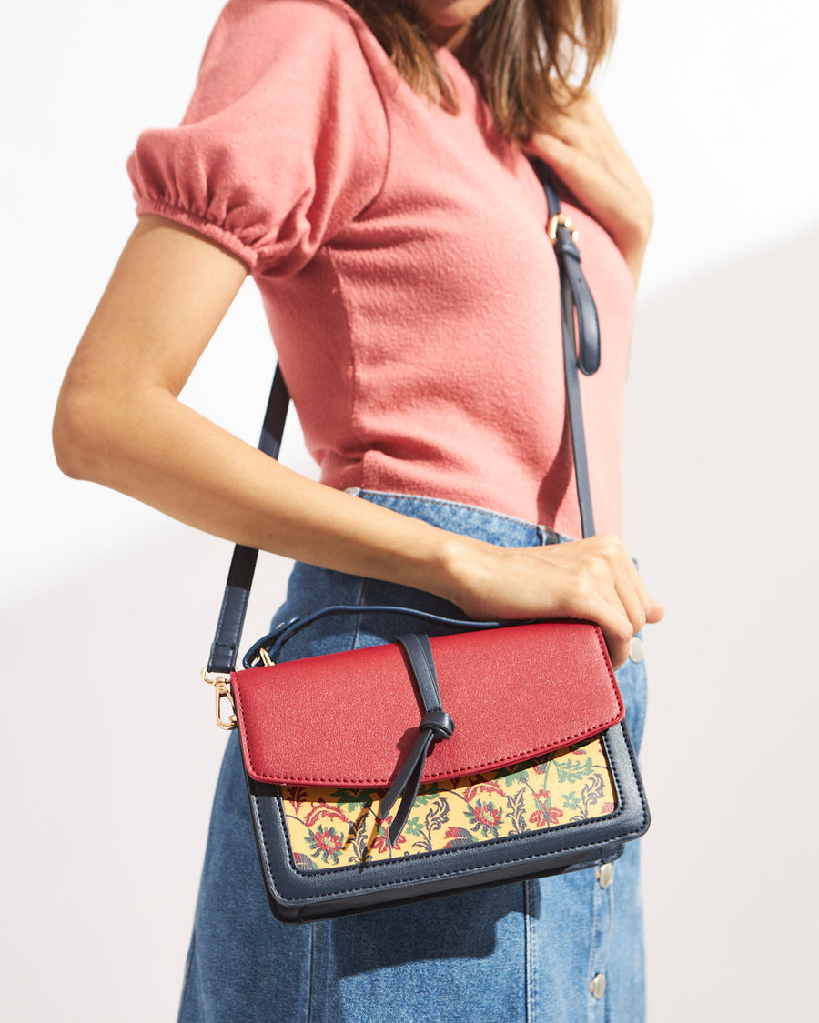 Woman wearing an elegant red and navy satchel bag with floral embroidery and a decorative knot on the front flap, paired with a pink top and denim skirt.