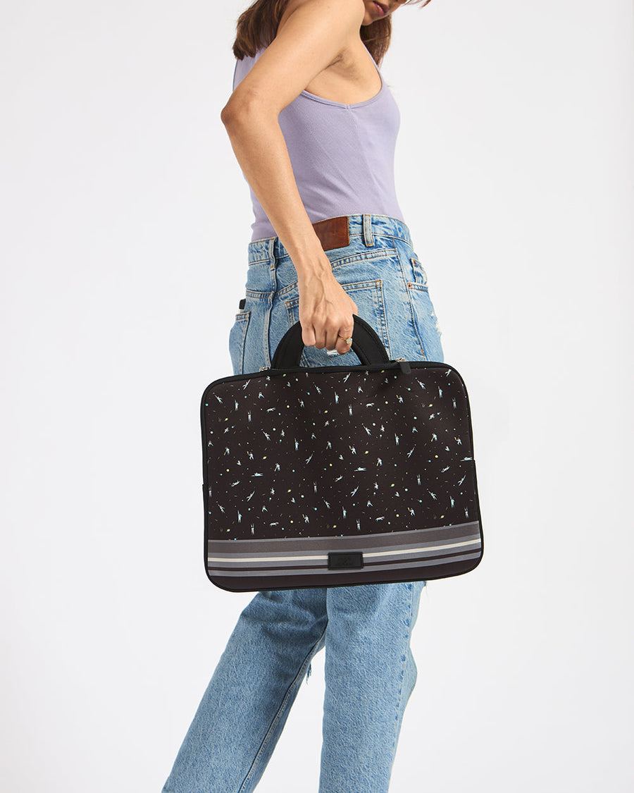 Woman in light purple tank top and blue jeans holding a black embroidered Milky Way laptop sleeve with space-themed embroidery and carrying handles.