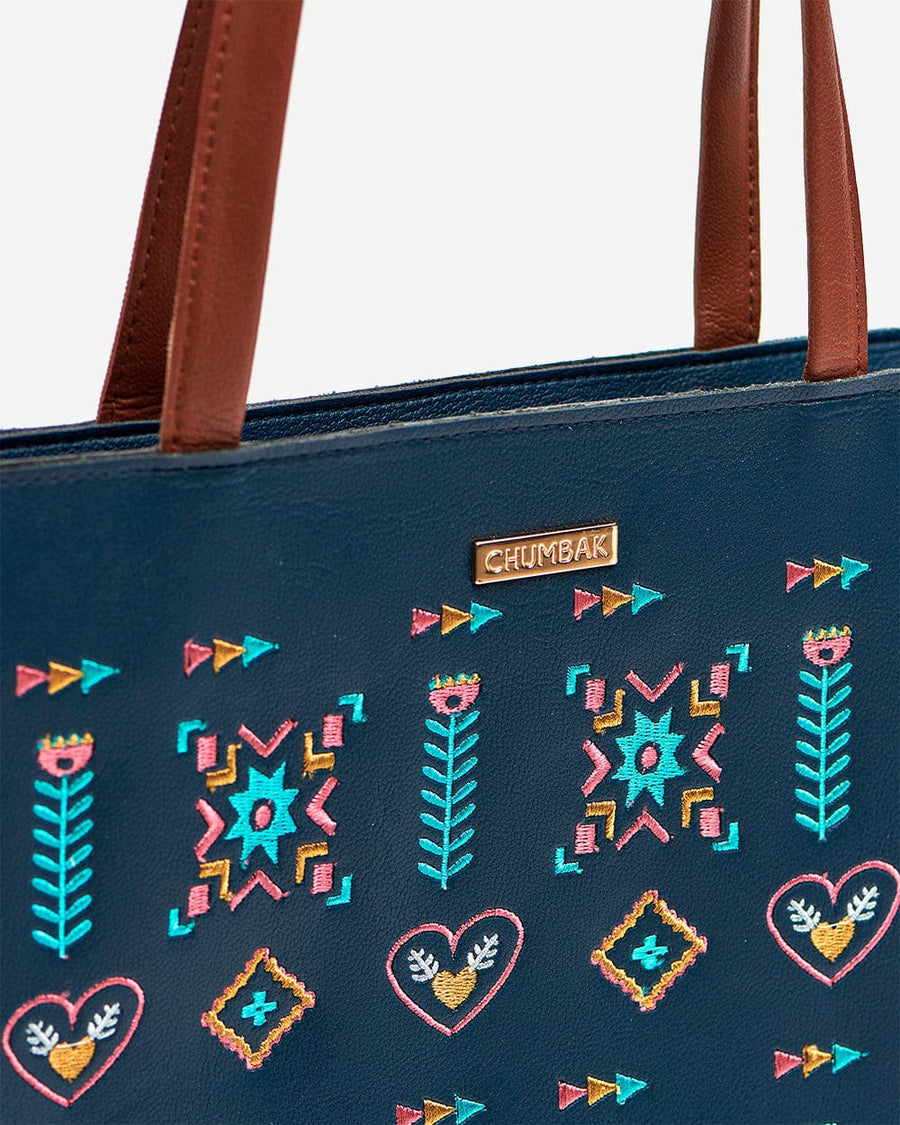 Close-up of intricate multicolor aztec embroidery detail on navy tote bag with a small gold brand badge
