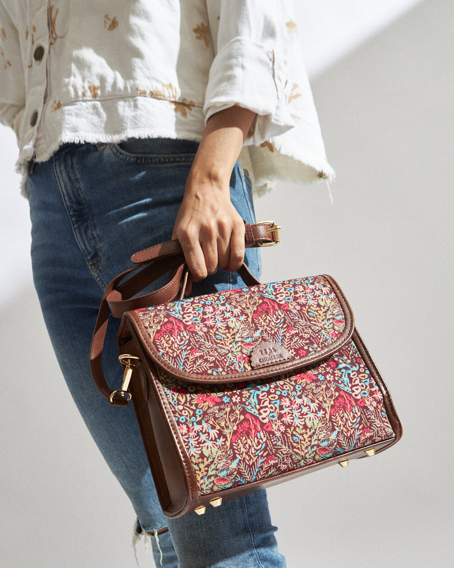 Woman holding a floral print crossbody bag with brown leather trim and adjustable strap, wearing blue jeans and a white floral jacket.