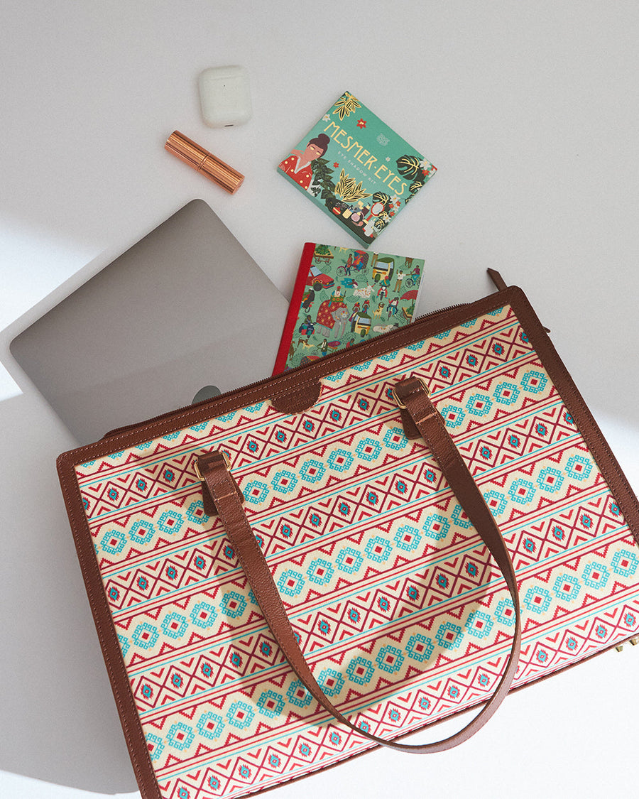 Aztec patterned office tote laid flat on a white surface beside a laptop, lipstick, AirPods case, and colorful notebook